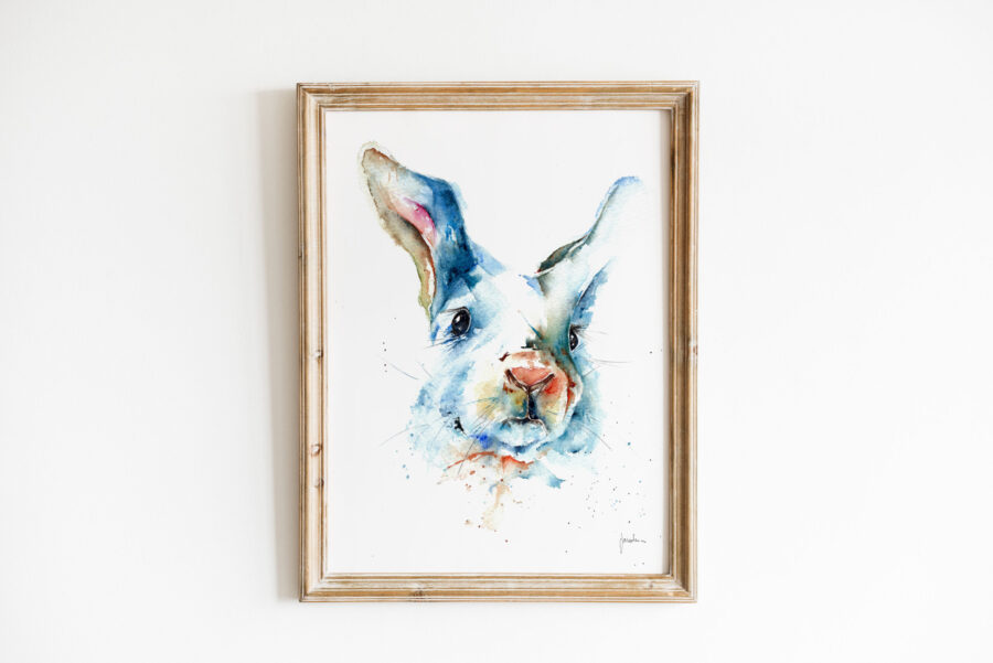 Cuadro enmarcado con una pintura en acuarela de un conejo con tonos azulados y detalles en rosa y naranja, colgado en una pared blanca con un marco de madera rústico.