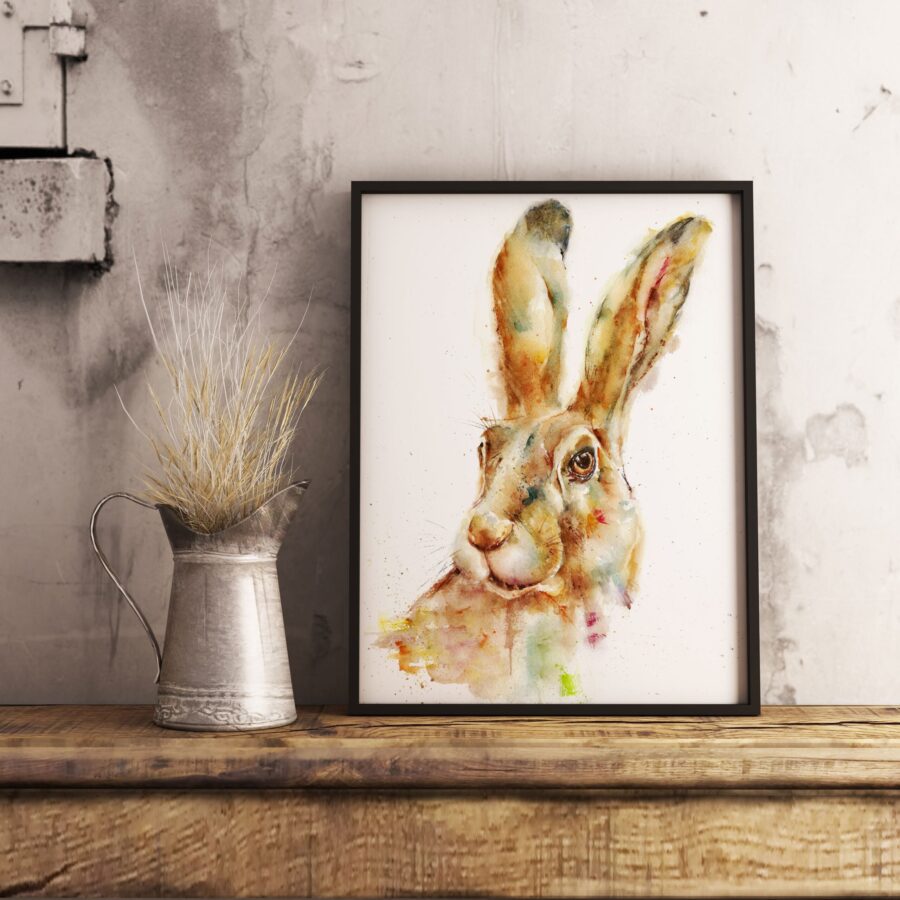 Pintura en acuarela de un conejo de orejas erguidas enmarcado, expuesto sobre una mesa de madera junto a un jarrón metálico con espigas secas