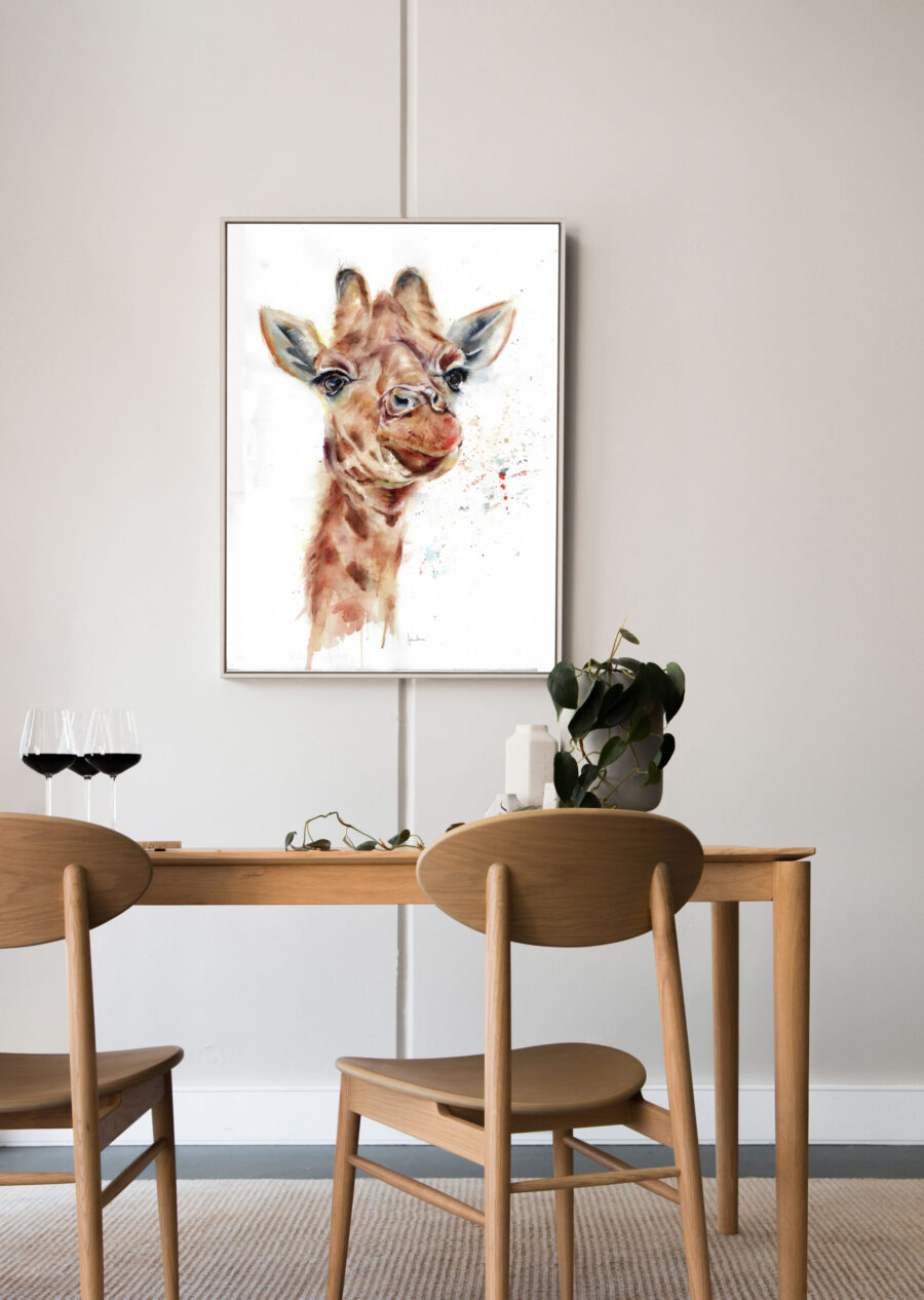 Cuadro con ilustración en acuarela de una jirafa, colgado en una pared sobre una mesa de comedor de madera con sillas a juego y copas de vino